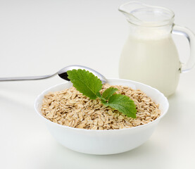 Milk and white bowl with dry oatmeal on a white background, breakfast