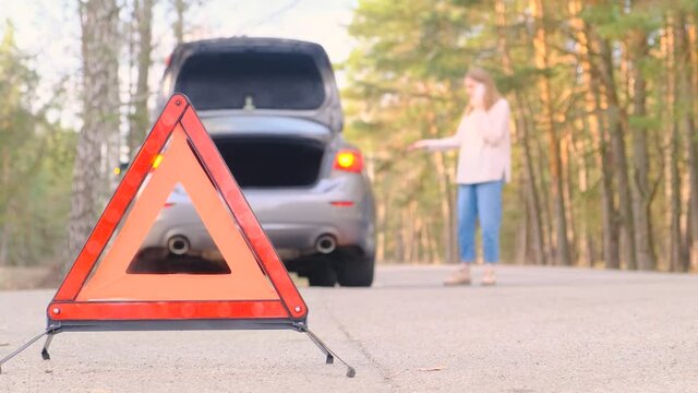 Car breakdown. A young woman walks along the road along the woods next to a broken car talking on the phone. Close-up of the emergency triangle, a woman in defocus. Car alarms flash
