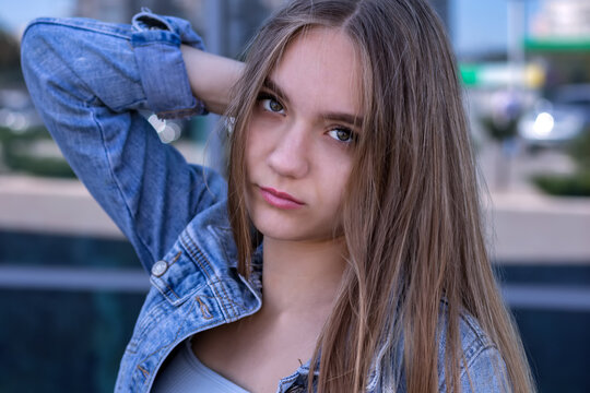 young beautiful charming stylish European girl, 16 years old, with long brown hair, brown eyes, wearing a denim jacket, street portrait of a modern teenage girl on a blue background.