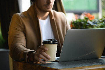 Cropped view of businessman in coat holding takeaway coffee near laptop in cafe 