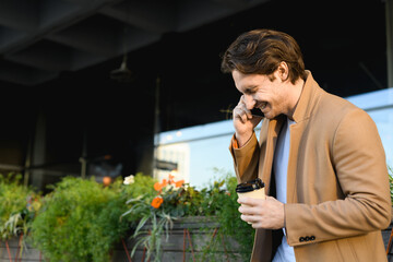 Side view of joyful businessman with takeaway coffee talking on smartphone on urban street 