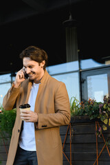 Good looking businessman in coat talking on smartphone and holding coffee to go on urban street 