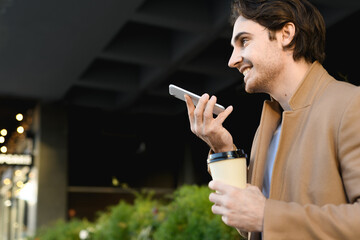 Smiling businessman in coat recording audio message on cellphone and holding takeaway drink on urban street 