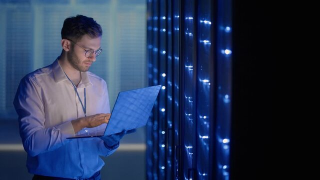 Male Server Engineer in Data Center. IT engineer inspecting a secure server cabinet using modern technology laptop coworking in data center.