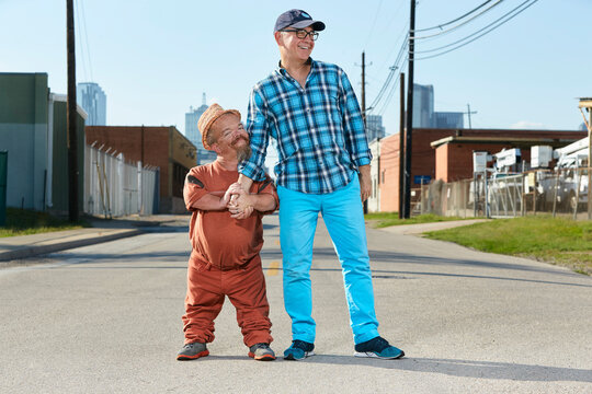Portrait of male dwarf little person hugging his friend, standing in middle of street. 