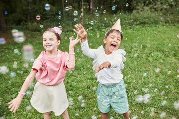 Portrait of two cute girls playing with bubbles while enjoying Birthday party outdoors in Summer - obrazy, fototapety, plakaty