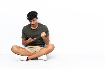 Young Argentinian man sitting on the floor surprised and sending a message