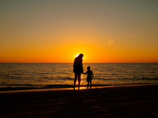 A young woman and a little girl stand on the sea beach and hold hands. In the background, the evening sun setting over the horizon.