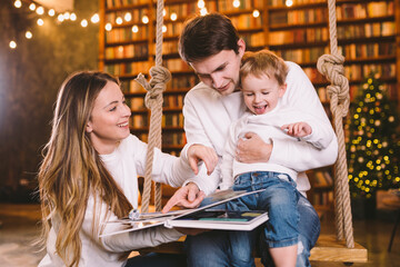 Family With Baby Relaxing On home Swing Seat in cozy dark living room on Christmas eve. Family...