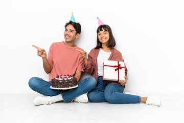 Young mixed race couple celebrating a birthday sitting on the floor isolated on white background pointing finger to the side in lateral position