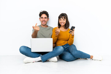 Young couple with a laptop and mobile sitting on the floor inviting to come with hand. Happy that you came