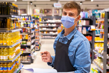 Young man working in a grocery store wearing protective medical mask for protection from virus over supermarket background holding in his hands the journal for inventory and the pen.
