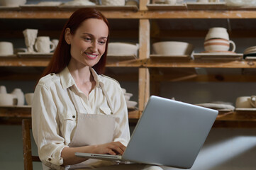 A young woman sitting at the laptop in a pottery workshop