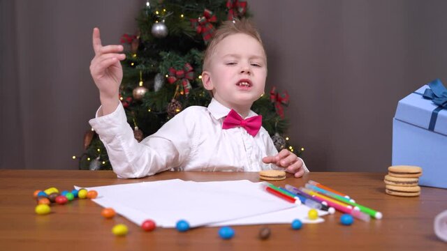 Spoiled boy does not want to draw postcard, write letter with wishes to Santa or does homework so he indulges, talks and eats candy, decorated Christmas tree on background.