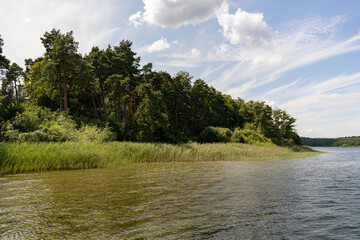 The shore line on a small lake. Green grass is growing in the area. A small forest is in the background. A white ship is on the water. Fluffy clouds are on the blue sky in this idyllic landscape.