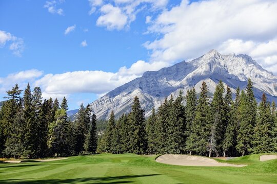 A beautiful view of a par 4 golf hole on a course with a huge mountain in the background, surrounded by forest, on a beautiful sunny day with blue sky, in the rocky mountains near Banff, Alberta