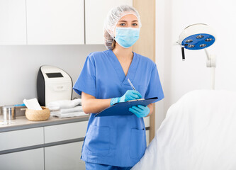 Portrait of confident professional female doctor in face mask standing in modern aesthetic clinic