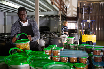 Focused African-American man working in food producing factory, arranging plastic buckets with...