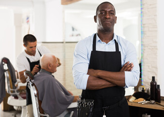 Confident African-American man hairdresser posing with folded arms in modern barber shop..