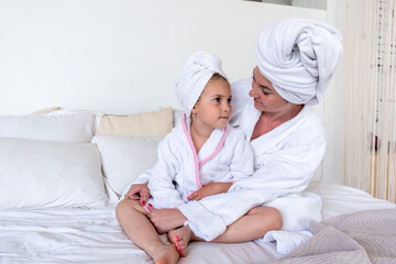 Mom and daughter are dressed in white terry robes hugging while sitting on the bed. Nursing communication within the family.