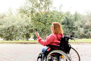 Fototapeta na wymiar Disabled teenage girl in a wheelchair takes a selfie while walking in the park. Side view. Full-fledged and a vibrant life for people with disabilities. Online communication. 