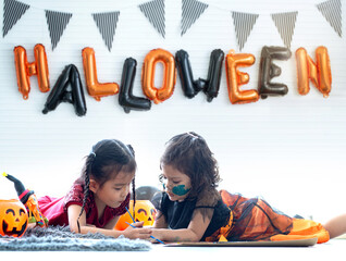 Little girl in Halloween costumes playing on floor, decorative flags and black and orange balloons in a Halloween theme on white wall, Halloween party concept