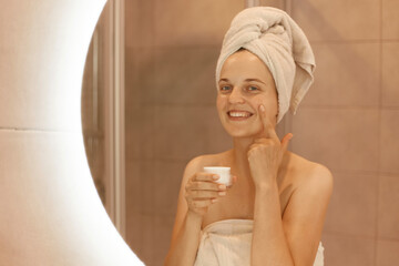 Indoor shot of happy smiling woman standing in front of mirror rubbing cosmetic cream on her face, putting on moisturizer on her facial skin in bathroom.