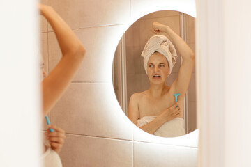 Indoor shot of unhappy woman shaving armpit in bathroom with razor, looking at her mirror...