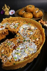 Rajasthani Ghevar. Ghewar is an Indian crunchy sweet dish made using refined flour, sugar, and ghee. Garnished with silver foil and dry fruits. Over dark background with copy space.