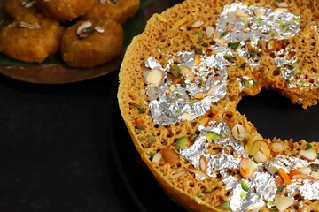 Rajasthani Ghevar. Ghewar is an Indian crunchy sweet dish made using refined flour, sugar, and ghee. Garnished with silver foil and dry fruits. Over dark background with copy space.
