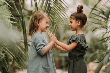 Two cute smiling little girls belonging to different races, in linen clothes, holding hands and...