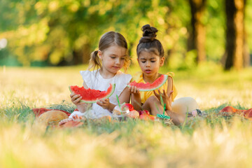 Two little mixed race kids consist of european and caucasian girls smiling with happiness, fun amusement, playing, sitting for picnic and eating piece of watermelon fruit in outdoor garden