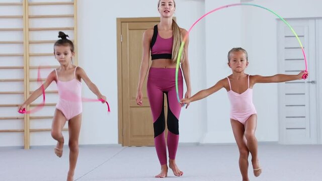 Two little girls gymnasts walking forwards while jumping over the rope and their female trainer watching and following them