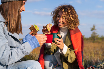 Two cheerful young girls having picnic outdoors during sunny autumn day. Happy girlfriends spend fall weekend in countryside eating sandwiches and drinking hot tea or coffee with happy relaxed smiles