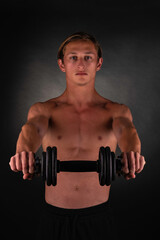 Fototapeta na wymiar Young athlete holding his dumbbell in front of him with his both arms during his routine on black and grey blackground. 