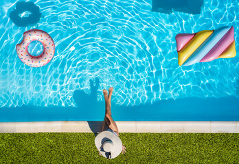 Aerial top down view of a woman with hat sitting at the swimming pool edge with colorful floats in...