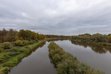 Fototapeta na wymiar Autumn landscape with a river. Cloudy autumn day by the river. View of the river with trees and bushes on the bank. Bright colors of autumn on a cloudy day.