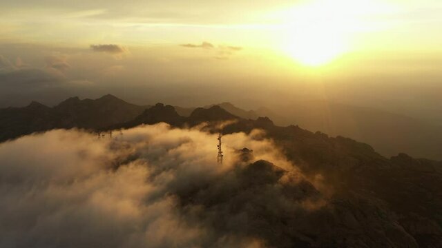 View from above, flying above the clouds, stunning aerial view of a mountain range with an antenna farm surrounded by clouds during a beautiful sunrise. Mount Limbara (Monte Limbara) Sardinia, Italy.