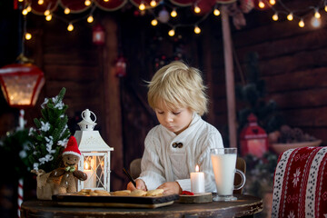 Beautiful toddler child, blond boy, writing letter to Santa Claus and eating cookies at home, cozy atmosphere