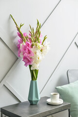 Vase with beautiful gladiolus flowers and cup of coffee on table in room