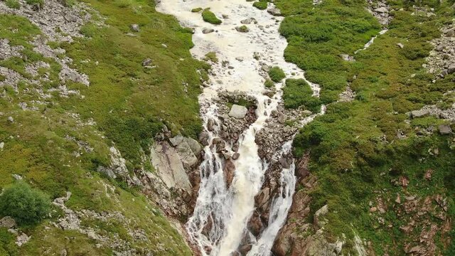 Long waterfall falls down along sharp rocks between steep cliffs with bushes and grass on top from mountain height aerial view