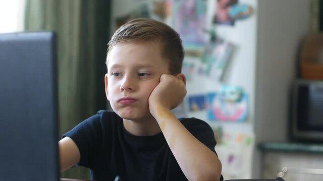 A 6-8 year old Caucasian boy working at a computer is very tired of his studies. Upset boy sighs heavily from home schooling at the laptop. Distance learning student