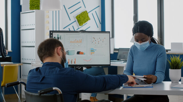 Invalid paralysed handicapped office manager explaining to black colleague financial startegy pointing graphs on clipboard wearing face masks against covid19 pandemic in new normal company