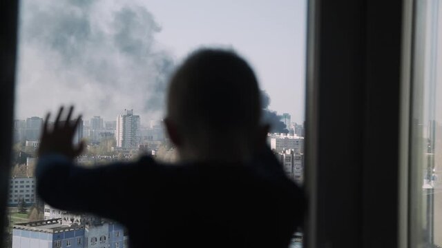 Silhouette of small child. Kid stands with his palms on the window and looks at the fire in city. Black smoke rises into the sky above high-rise buildings. Boy is worried. Child safety.