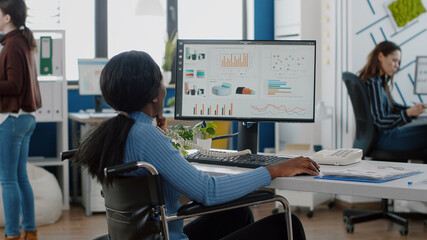 Busy paralysed handicapped black employee answering phone talking with colleague sitting immobilized in wheelchair in start up business office resolving financial problems discussing statistics graphs