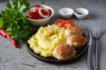 A hearty lunch: mashed potatoes, pork cutlets, tomato slices in a plate, tomato salad in a white bowl, fresh greens on a gray background. Close-up