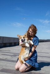 A girl with long red hair in a blue dress hugs her corgi dog in the park, in the afternoon, on the beach. Friendship, vaccination of dogs, morning walk, rest, domestication