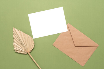 Blank card mockup, craft envelope and dry palm leaf on olive color paper background. Top view, flat lay. Stationery modern still life.