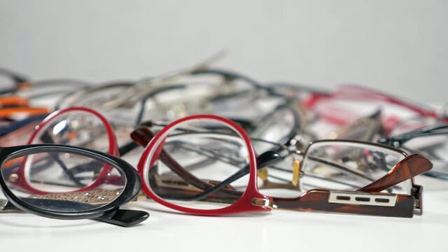 Camera shows pile of eyesight glasses with bright multi-colored frames lying on white wooden table against grey wall extreme closeup
