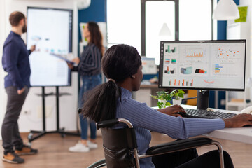 Handicapped african american businesswoman in wheelchair analyzing financial statistics on computer. Paralyzed disabled woman working at company strategy in startup business corporate office